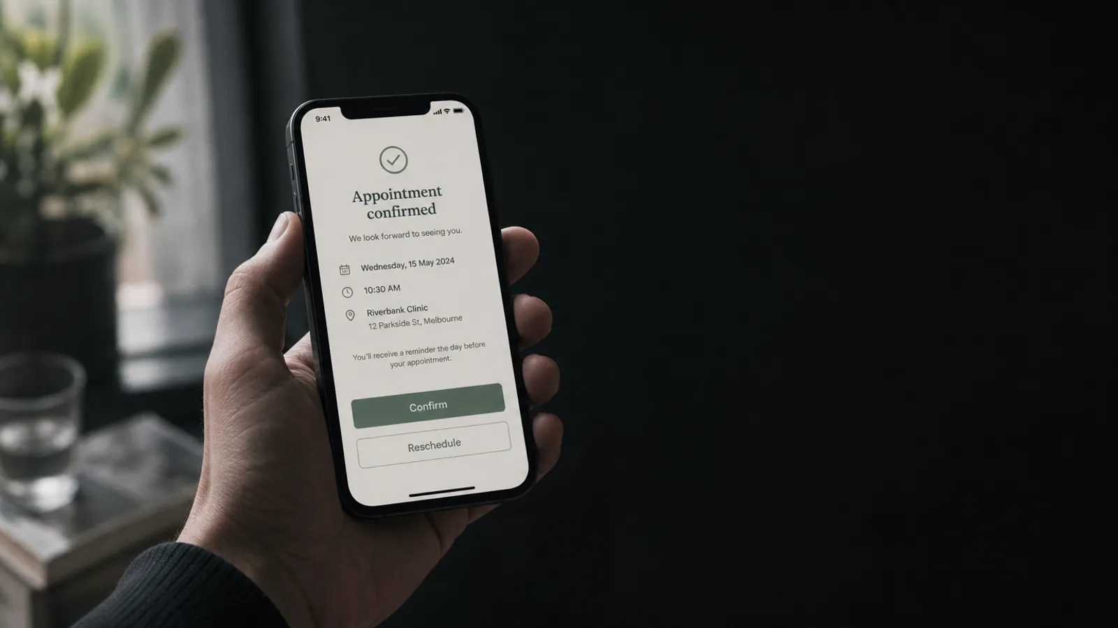 Hand holding a phone showing an appointment reminders confirmation message on a calm dark clinic desk in soft light