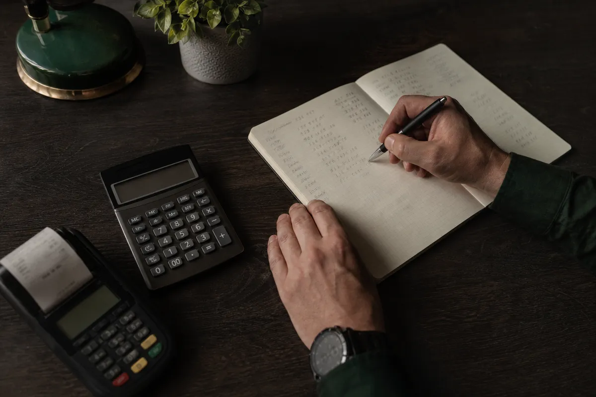 Owner's hands working through voucher figures in a notebook beside a calculator on a dark desk