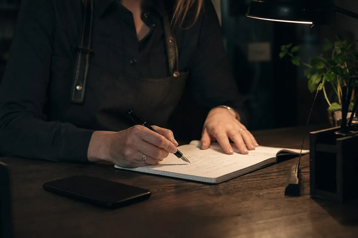 Hands writing short text message drafts on grid paper with a pen, a phone face down beside the notebook