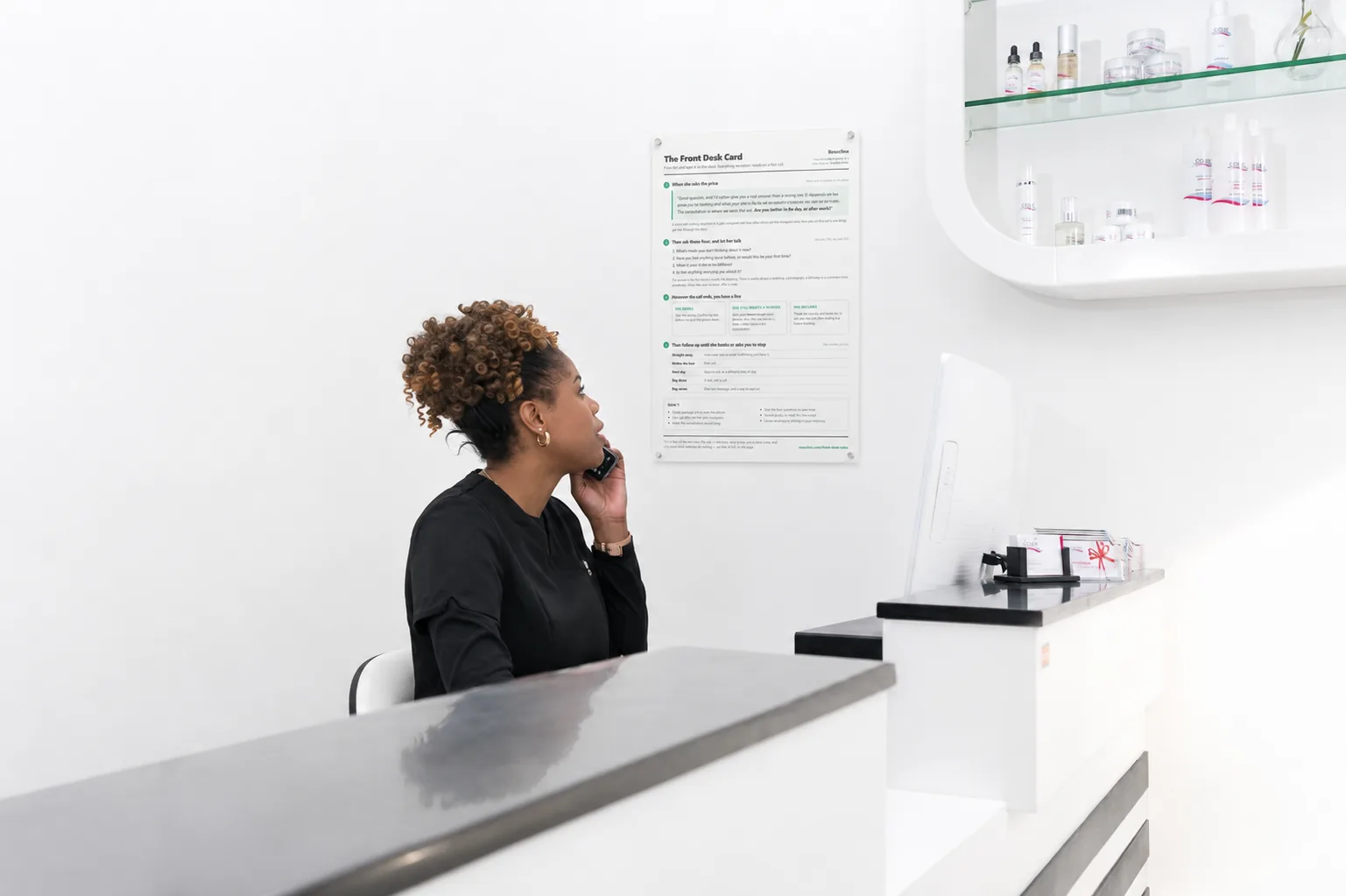A clinic receptionist on the phone, with the printed Front Desk Card on the wall behind her.